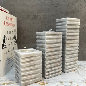 May include: Three white marble pillar candles of varying sizes. The candles are stacked in a graduated pattern, with the largest candle in the back and the smallest candle in the front. The candles are on a white marble surface.