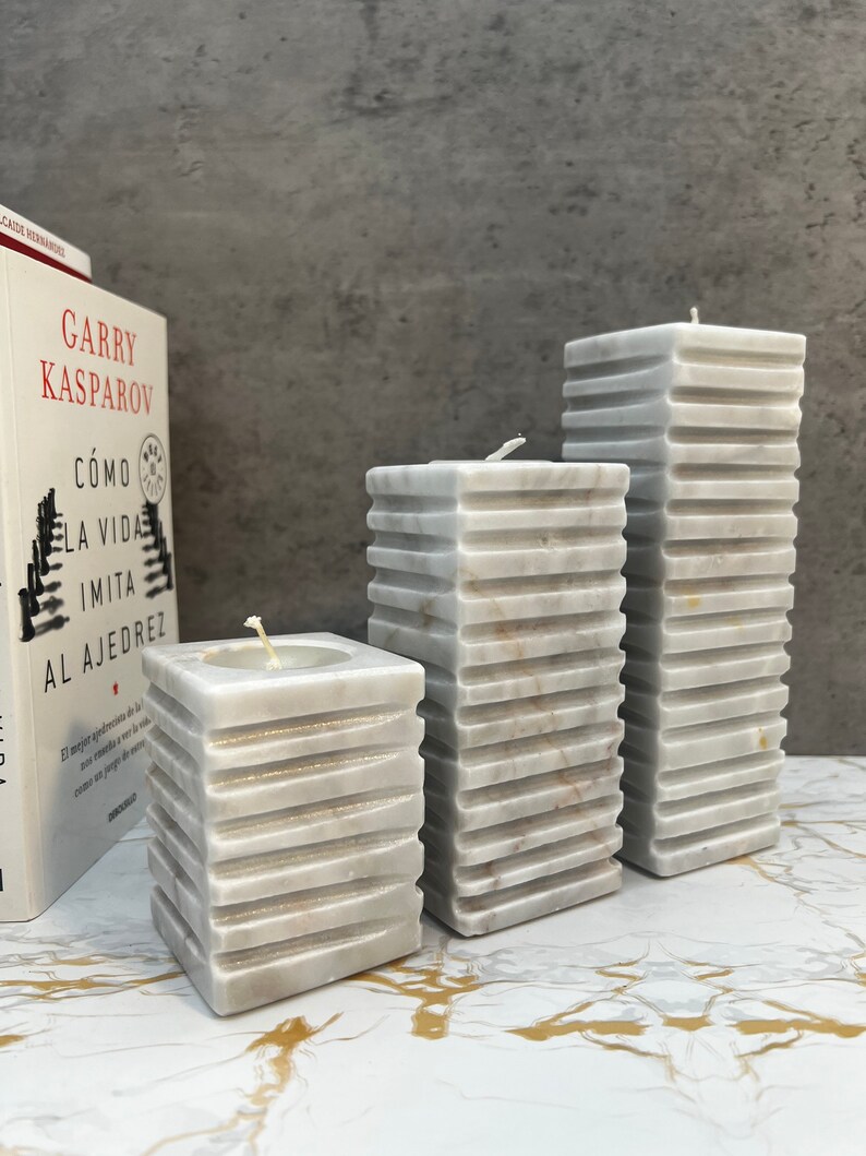 May include: Three white marble pillar candles of varying sizes. The candles are stacked in a graduated pattern, with the largest candle in the back and the smallest candle in the front. The candles are on a white marble surface.