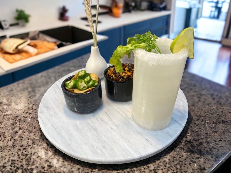 May include: A white marble serving tray with a clear glass filled with a white drink, a small bowl of green jalapenos and chips, and a small bowl of brown and black food. The drink has a salted rim and a lime wedge garnish.