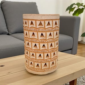 Könnte beinhalten: Eine zylindrische, beige Keramikvase mit einem geometrischen Muster aus Dreiecken und Quadraten. Die Vase steht auf einem hellen Holztisch, im Hintergrund ein graues Sofa. Die Vase ist etwa 25 cm hoch.