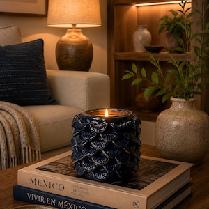 Puede incluir: Una vela azul marino encendida en un portavelas decorativo con un diseño en forma de escamas. La vela está sobre dos libros con los títulos "MÉXICO" y "VIVIR EN MÉXICO". Una lámpara encendida y objetos decorativos están en el fondo.