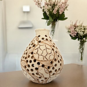 May include: A white ceramic candle holder with a decorative design of stylized birds and leaves. The holder has a round shape and is perforated with small holes.