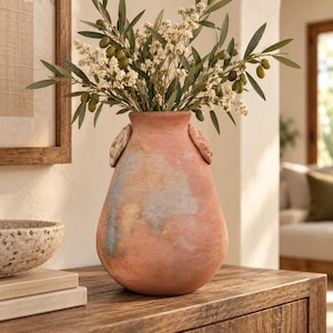 May include: A terracotta vase with a rounded body and two small side handles, featuring a weathered finish with muted patches of blue and grey. The vase is filled with a bouquet of green olive branches and small white flowers.