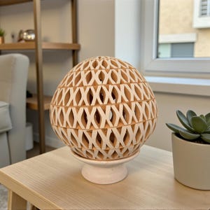 Op de afbeelding: Een decoratieve, bolvormige lamp met een roosterontwerp, gemaakt van lichtbruin materiaal, staat op een witte sokkel. De lamp staat op een lichte houten tafel naast een kleine vetplant in een pot. De kamer heeft een neutraal kleurenpalet.