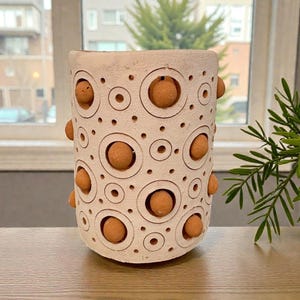 May include: A cylindrical, off-white ceramic vase with a textured surface. The vase features a pattern of circles and raised, spherical accents in a warm, earthy tone. The design is repeated around the vase. A decorative item for the home.