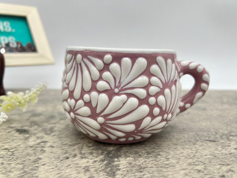 May include: A pink ceramic mug with a white floral design. The mug has a rounded shape and a handle.