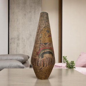 May include: A tall, decorative vase with a textured, speckled brown surface. The vase features a painted design with a face, a rainbow, and vertical lines in red, black, and green. The vase is displayed on a light-colored table.