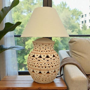May include: A beige ceramic table lamp with a cream-colored lampshade. The lamp base has a carved geometric design. The lamp is on a wooden table next to a houseplant and a sofa.