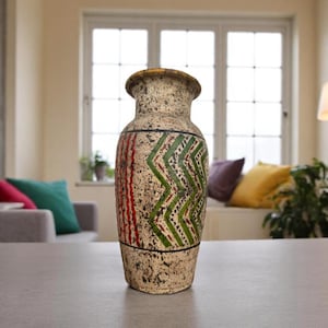 May include: A decorative, textured vase with a cream-colored base and a patterned design. The vase features vertical red stripes and green zigzag patterns. The top rim is a golden yellow color. The vase is a decorative home accent.
