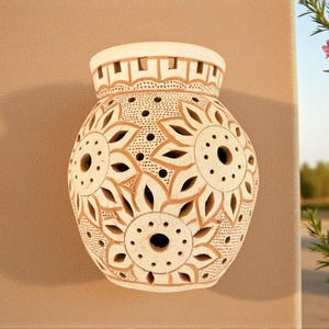 Op de afbeelding: Crèmekleurige keramische vaas met ingewikkelde bloemvormige uitgesneden ontwerpen. De vaas heeft een afgeronde body en een licht uitlopende rand. De uitsparingen creëren een patroon van bloemen en bladeren, met kleine cirkelvormige gaten. De vaas is ongeveer 25 cm hoog.
