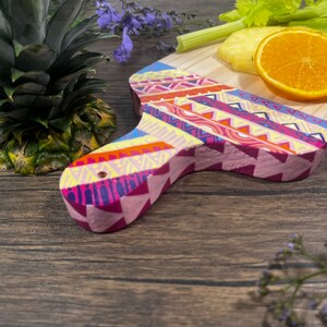 Pu&ograve; includere: Un tagliere di legno colorato con un motivo geometrico in tonalit&agrave; di rosa, giallo, blu e arancione. Il tagliere ha un manico ed &egrave; decorato con un motivo tribale.
