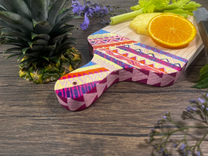 Pu&ograve; includere: Un tagliere di legno colorato con un motivo geometrico in tonalit&agrave; di rosa, giallo, blu e arancione. Il tagliere ha un manico ed &egrave; decorato con un motivo tribale.