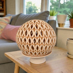 Op de afbeelding: Een decoratieve, bolvormige, crèmekleurige keramische lamp met een rasterontwerp. De lamp staat op een witte voet. Het uitgesneden patroon laat licht door. Woondecoratie.