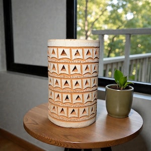 Op de afbeelding: Een cilindervormige, crèmekleurige keramische vaas met een herhalend patroon van uitgesneden driehoeken en vierkanten. De vaas is ongeveer 25 cm hoog en staat op een ronde houten tafel. Een kleine groene plant in een pot staat naast de vaas.