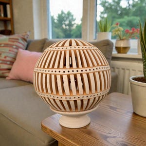 Op de afbeelding: Een decoratieve, bolvormige lamp met een witte voet en een patroon met opengewerkt design. De lamp is gemaakt van een lichtbruin materiaal met verticale uitsparingen en kleine gestippelde gaten. Hij staat op een houten tafel, met een bank en planten op de achtergrond.