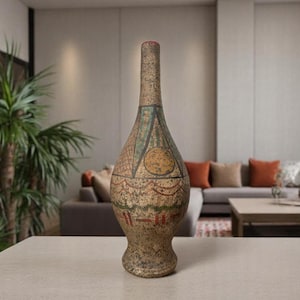 May include: A tall, decorative vase with a narrow neck and bulbous base. The vase is painted with geometric patterns in shades of brown, green, and red. It stands on a light-colored surface, with a blurred background of a living room.