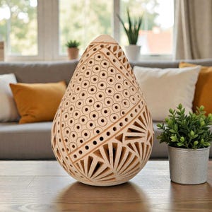 Op de afbeelding: Een beige, druppelvormige decoratieve lamp met ingewikkelde uitgesneden patronen. De lamp heeft geometrische ontwerpen en cirkelvormige perforaties. Hij staat op een houten tafel, met een kleine plant in een pot in de buurt.