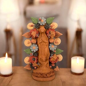 May include: A terracotta-colored religious figurine of a woman with hands clasped in prayer, adorned with colorful clay flowers and leaves. The figurine is surrounded by lit candles, creating a warm and inviting ambiance. The flowers are in shades of red, yellow, blue, and green.