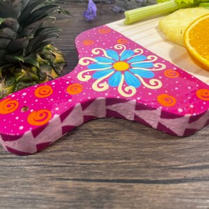 Pu&ograve; includere: Un tagliere in legno rosa con un design floreale dipinto a mano. Il tagliere ha un manico in legno chiaro e un motivo geometrico rosa e bianco sul fondo.