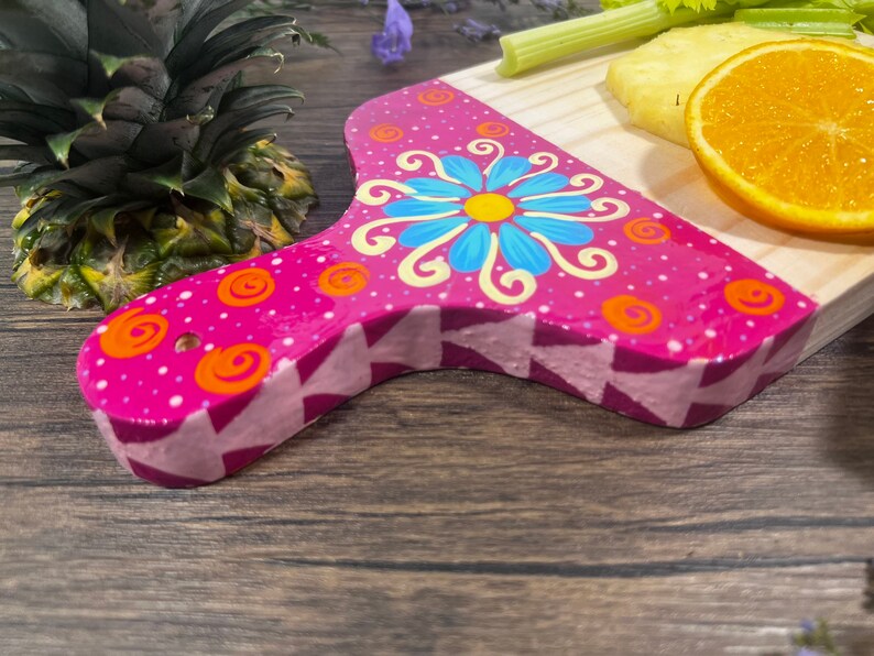 Pu&ograve; includere: Un tagliere in legno rosa con un design floreale dipinto a mano. Il tagliere ha un manico in legno chiaro e un motivo geometrico rosa e bianco sul fondo.
