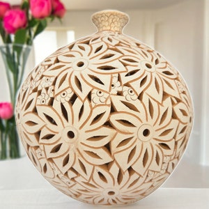 May include: A white ceramic vase with a round shape and intricate floral designs carved into the surface. The vase has a delicate, openwork pattern that creates a light and airy feel.