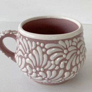 May include: A pink ceramic mug with a white floral design. The mug has a rounded shape and a handle.
