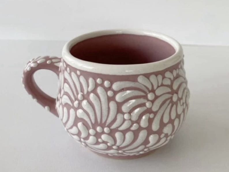 May include: A pink ceramic mug with a white floral design. The mug has a rounded shape and a handle.