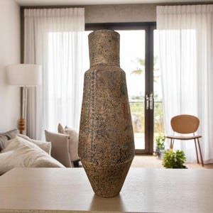 May include: A tall, textured, brown vase with a wide base and a narrow neck. The vase has a speckled appearance with hints of green and red. It stands on a light-colored surface in a well-lit room.