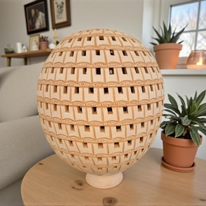 Op de afbeelding: Een bolvormige, beige keramische lamp met een herhalend vierkant patroon van uitgesneden ramen. De lamp staat op een kleine, ronde voet. De lamp staat op een houten tafel.