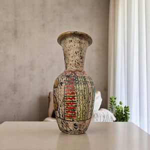 May include: A tall, decorative vase with a speckled, off-white finish. The vase features a painted design with red, green, and yellow accents. The vase is displayed on a white table in front of a neutral-toned wall.
