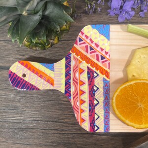 Pu&ograve; includere: Un tagliere di legno con un motivo geometrico colorato dipinto sul manico. Il tagliere &egrave; decorato con una fetta d'arancia e un pezzo di ananas.