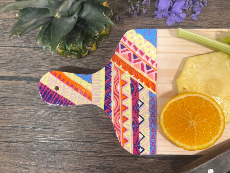 Pu&ograve; includere: Un tagliere di legno con un motivo geometrico colorato dipinto sul manico. Il tagliere &egrave; decorato con una fetta d'arancia e un pezzo di ananas.