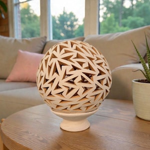 Op de afbeelding: Een decoratief, bolvormig object met een geometrisch uitgesneden ontwerp. De bol is gebroken wit en staat op een bijpassende sokkel. Het object staat op een houten tafel, met een bank en plant op de achtergrond. De bol meet ongeveer 25 cm in diameter.
