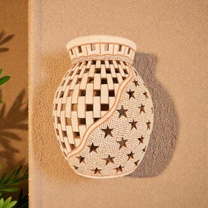Op de afbeelding: Crèmekleurige, decoratieve wandvaas met een geweven patroon en steruitsparingen. De vaas heeft een cilindrische vorm met een brede opening en is bevestigd aan een getextureerde, bruinbeige muur. Het ontwerp omvat een sterrenband en een geometrisch patroon.