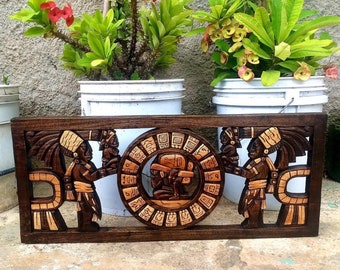 May include: A dark brown wooden wall hanging with a carved design of two figures holding a circular object with Mayan glyphs. The figures are wearing headdresses and traditional clothing. The background is a light brown wood.