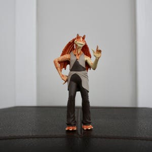 May include: A detailed figurine of a character with orange skin, large ears, and a long, red headpiece. The figure is wearing a gray and black outfit and is posed with one hand raised. The figure is standing on a dark surface.