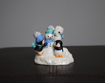 May include: A whimsical figurine featuring a snowman and three animal figures. The snowman wears a blue and green hat and scarf. Two penguins and a polar bear are also present, all adorned with winter accessories. The base is a sparkly white mound.