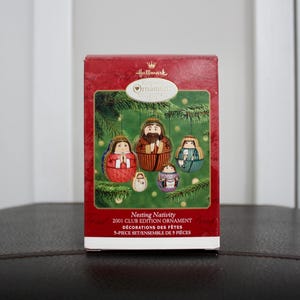 May include: A red Hallmark box featuring a nesting nativity set. The box has a green background with illustrations of the nativity figures. Text on the box reads "Nesting Nativity 2001 Club Edition Ornament".