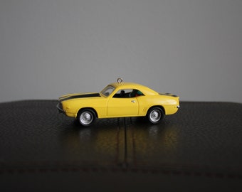 May include: A yellow toy car with black racing stripes on the hood. The classic car has a detailed design, including wheels and a small loop for hanging. The car is a miniature replica of a vintage model.
