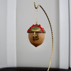 May include: A decorative acorn ornament with a red bow and a gold-colored metal hook. The acorn has a brown, textured body and a detailed cap. The ornament is suspended from a gold-colored metal stand.