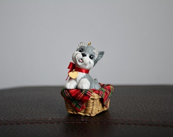 Può includere: Statuetta di cucciolo grigio e bianco con un nastro rosso e un'etichetta a forma di cuore dorata, seduta in un cestino intrecciato foderato con tessuto a quadri rosso e verde. Il cucciolo ha un'espressione felice. Un oggetto decorativo.