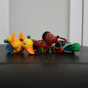 May include: A row of colorful pull-along toys: a yellow duck, a brown dog, and a green turtle. Each toy has wheels and is connected by a red or green string. The toys are on a dark surface.
