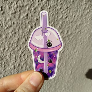Puede incluir: Una pegatina con una taza de t&eacute; boba de dibujos animados con un dise&ntilde;o de galaxia. La taza es morada y rosa, con una pajita y una tapa. El contenido de la taza incluye una luna creciente, estrellas y peque&ntilde;as criaturas negras.