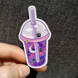 Puede incluir: Una pegatina con un dise&ntilde;o de t&eacute; de burbujas morado con un interior con tem&aacute;tica de galaxia. La pegatina incluye una pajita, nubes, estrellas, una luna creciente y peque&ntilde;os duendes de holl&iacute;n negros. La taza tiene un borde rosa y la sostiene una mano.