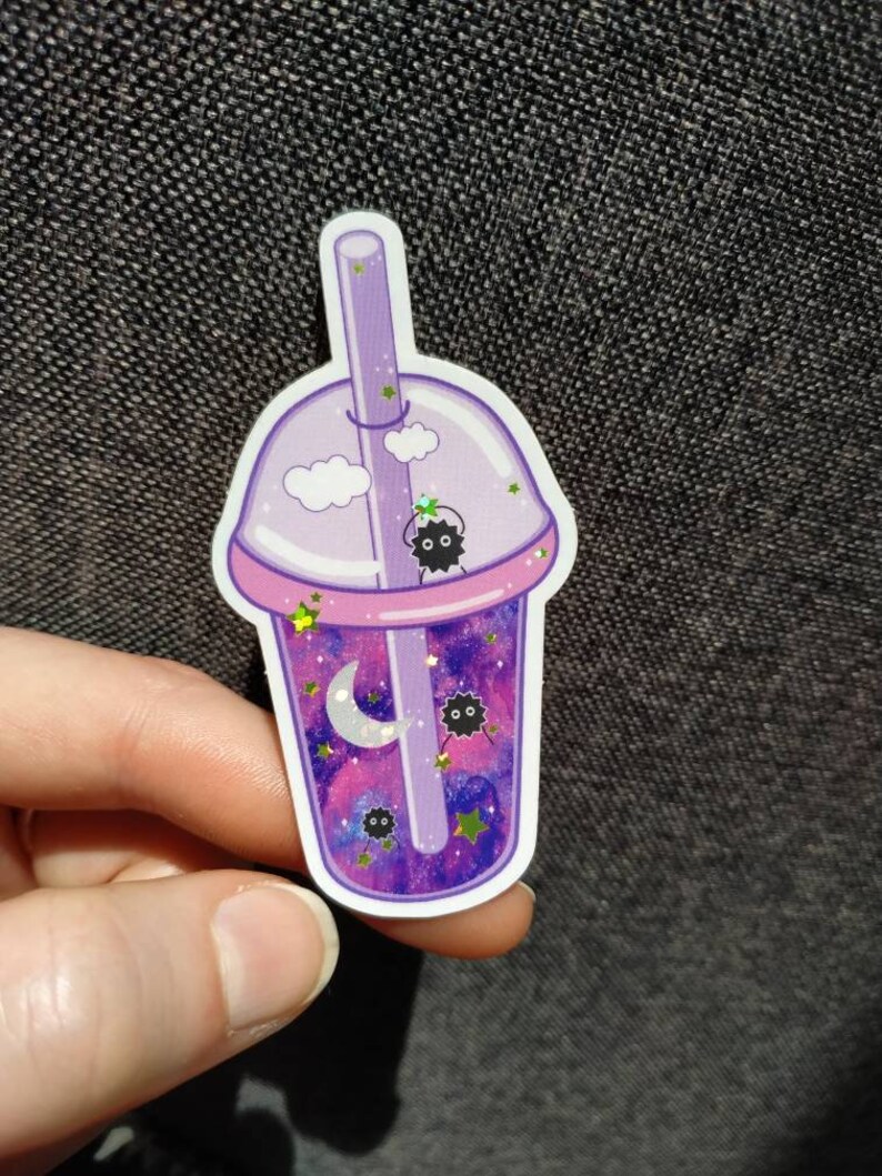 Puede incluir: Una pegatina con un dise&ntilde;o de t&eacute; de burbujas morado con un interior con tem&aacute;tica de galaxia. La pegatina incluye una pajita, nubes, estrellas, una luna creciente y peque&ntilde;os duendes de holl&iacute;n negros. La taza tiene un borde rosa y la sostiene una mano.