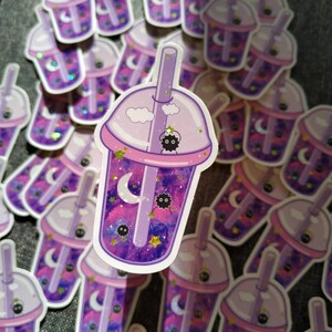 Puede incluir: Una colecci&oacute;n de pegatinas con un dise&ntilde;o de t&eacute; de burbujas morado. La taza de t&eacute; de burbujas tiene un interior con tem&aacute;tica de galaxia con una luna creciente, estrellas y un duende de holl&iacute;n negro. La taza est&aacute; rematada con una tapa y una pajita de color p&uacute;rpura claro.