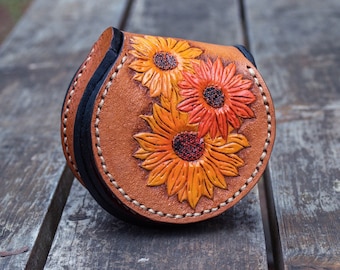 May include: A brown leather coin purse with a stitched edge and a floral design featuring three orange and yellow sunflowers.