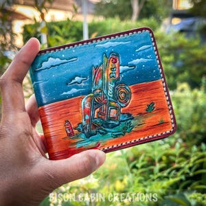 Puede incluir: Cartera de cuero hecha a mano con una escena desértica con una gasolinera vintage bajo un cielo azul. La cartera tiene un borde cosido y el texto "Cool 360". La parte inferior de la cartera tiene el texto "Bison Cabin Creations".