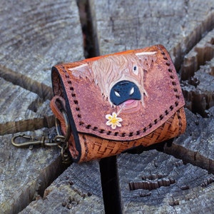 Könnte beinhalten: Ein brauner Leder-Schlüsselanhänger mit einem detaillierten Bild einer Highland-Kuh mit einer Blume im Maul. Der Schlüsselanhänger hat einen Metallring und ein genähtes Design.