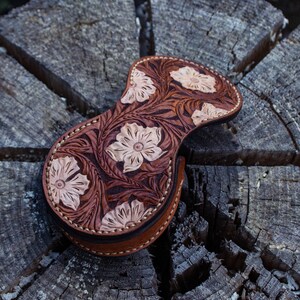 A brown leather pouch with a floral design. The pouch is decorated with a carved floral pattern in a light brown color. The coin purse is closed with a button .